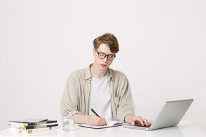 Portrait of serious young man student wears beige shirt and spectacles writing and study at the table using laptop computer and notebooks isolated over white background photo