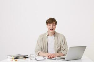 Portrait of amazed young man student wears beige shirt looks surprised and study at the table with laptop computer and notebooks isolated over white background photo