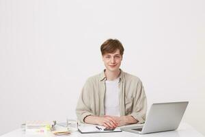 retrato de sonriente atractivo joven masculino estudiante usa beige camisa trabajando a el mesa con ordenador portátil computadora y cuadernos y estudiar aislado terminado blanco antecedentes foto
