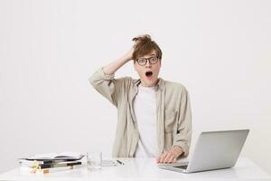 Closeup of shocked young man student with opened mouth wears spectacles and beige shirt looks amazed working with laptop computer and notebooks at the table isolated over white background photo