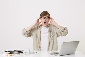 Portrait of astonished young man student with opened mouth wears glasses and beige shirt looks shocked sitting at the table with laptop computer and notebooks isolated over white background photo