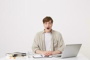 Portrait of shocked young man student opened mouth wears beige shirt feels astonished and using with laptop computer and notebooks at the table isolated over white background photo