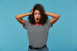 Shocked African American female stares at camera and keeps mouth opened, scratches head and looks surprisingly, realizes bad news, stands against blue background. People, reaction, ethnicity photo