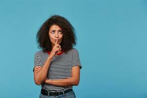 joven afro americano mujer con afro peinado vistiendo despojado camiseta terminado aislado azul antecedentes con Copiar espacio pensando reflexionando acerca de alguna cosa concentrado con dedo en labios. foto