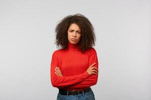 Serious suspicious latino woman with curly afro hair standing with crossed hands and frowning, expressing distrustfulness and disappointment, over white background photo
