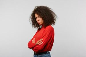 ofendido trastornado latino mujer con afro peinado en un rojo manga larga en pie oblicuo con manos cruzado en un blanco antecedentes foto