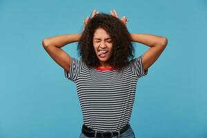 Close up of funny joking beautiful young woman, feels happy, makes horns on her head with fingers, shows tongue, closed eyes, isolated over blue background photo