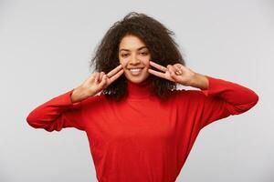 Close up of an happy young girl showing peace gesture. African American smiling woman with afro hairstyle looking at camera and doing victory signs with fingers. Isolated over white background photo