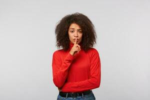 bonito joven mujer con afro peinado teniendo secreto mientras participación dedo en labios y demostración silencio firmar en pie en blanco antecedentes foto