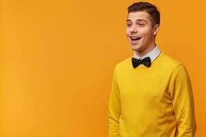 Joyful sociable excited attractive guy, smartly dressed in a yellow sweater with bow-tie, looks happy smiling,standing sideways looking towards empty space to place your advertisement, isolated photo