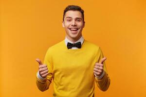 Portrait of attractive man with thumbs up looks like has a success, smartly dressed in yellow sweater over white shirt with bow-tie, happy excited face expression, showing positive gesture, isolated photo