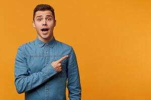 Man in panic nervous excited dressed in denim shirt pointing right with his finger, feals fear, isolated on yellow background with copy space for your text or advertisement photo