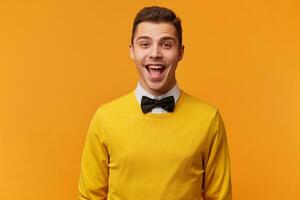 Studio shot of excited funny young attractive man, smartly dressed in a yellow sweater with bow-tie, has happy joyful face expression, smiling, isolated on yellow background photo