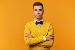 Young businessman with serious face expression ponder on question, concentrated on some idea, thoughtfully stands with arms crossed, dressed in yellow sweater and bow-tie,yellow background photo