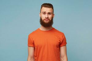 An adult guy with a thick beard wearing a red T-shirt isolated against a blue background feels distressed upset, with insult sad expression on the face, is ready to cry from injustice photo