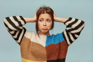 Close up of emotional woman with a sad expression on face, crooked lower lip with a disgruntled look. The girl feels disappointed looks left side keeps head with hands, isolated over blue background photo