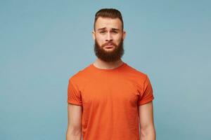 Sad crying bearded guy wearing a red T-shirt isolated against a blue background feels distressed upset, with insult sorrowful expression on the face, is ready to cry from injustice photo