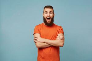 Emotional happy excited funny man with a heavy beard stands with arms crossed and opened mouth in surprisement dressed in red t-shirt isolated over blue background shows wow expression photo