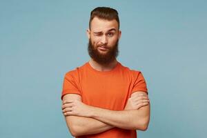 A young guy with a beautiful thick beard dressed in a red T-shirt isolated on a blue background, standing with arms crossed looking slightly dismissively arrogant with disapproval, one eye is closed. photo