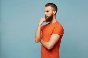 Young attractive strong cleaver man with a fashionable haircut thick beard standing half turn to camera dressed in a red T-shirt holds his fist near chin reflects on something thinks philosophizes. photo