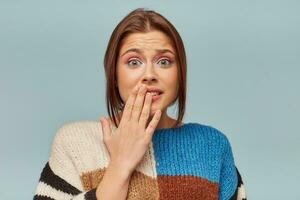 Confused young girl shrugging shoulders, having guilty look, feeling sorry for doing something wrong and making terrible mistake, keeps hand on lips, over blue background photo