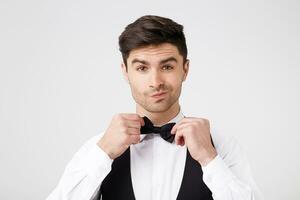 Self-confident brunet dressed in a smart suit straightens black butterfly has no doubt that he looks great and the date will go superbly, isolated white studio background photo