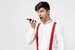 Furious handsome man is screaming on someone via telephone conversation. Guy keeps phone looks at it and yelling with wide opened mouth,wears white shirt and red suspender photo