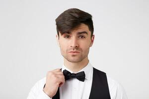 Attractive handsome smartly dressed dark-haired guy corrects his bow tie,looks charming confident inviting, isolated over white background photo