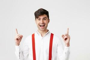 Indoor photo of happy glad handsome guy,looks amazed excited,dressed in white shirt and red suspenders, points with index fingers upwards on a blank white wall with copy space your promotional content