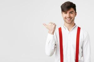 cerca arriba de elegante atractivo chico en blanco camisa y rojo liga señalando aparte con agradable sonrisa anunciar alguna cosa genial fantástico, en pie terminado blanco pared foto