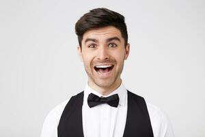 The stylish man fascinated and excited. Close up of the amazed attractive young male in black bowtie looks camera with a toothy surprised smile, standing over white wall photo