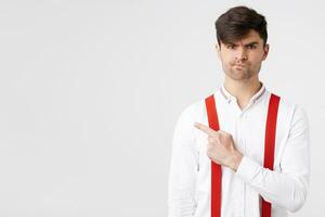 joven elegante atractivo chico en blanco camisa y rojo liga señalando con índice dedo a el izquierda lado con insatisfecho disgustado facial expresión hacer no Consejo algo, de pie terminado blanco pared foto