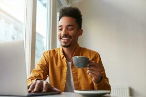 sitting at a table in a cafe and working on a laptop, wears in yellow shirt, drinks aromatic coffee, communicates with his sister which is far in another country , enjoys the work of a freelancer. photo