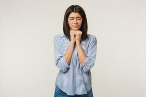 Girl folded hands in wishing gesture and eyes tightly closed while making a wish. Woman praying for close person, says thanks to God for help, wanting some present, hopes for luck, over white wall photo