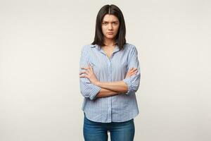 Studio shot of young brunette woman, frowing, unhappy, listens to someone with sadness, upset, standing with arms crossed dressed in blue jeans and striped shirt isolated over white background photo