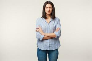 Indoor photo of brunette woman, looks with distrust wary suspicion, tense, listens to someone with doubt, stressful, standing with arms folded dressed in jeans and striped shirt, over white wall