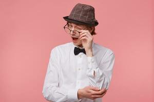 Attractive guy isolated on pink background, dressed in white shirt, hat and black bow tie pretends to lower the glasses and winks happily with approval, supports favourites, with joy and happiness photo