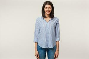 Studio photo of charming attractive smiling pleasant young brunette standing isolated over white background dressed in jeans and striped shirt.