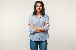 Young brunette woman, calm, attentive, listens with respect, pays attention, standing with arms crossed dressed in blue jeans and striped shirt isolated over white background photo