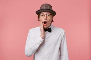 Shocked young guy dressed in white shirt, hat and black bowtie wears glasses, opened mouth widely and raised hand to a cheek in surprise, filled with emotion, isolated on pink background photo