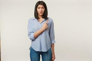 Worried nervous young brunette looks camera in panic, scared, mouth opened, pointing index finger to the right side on copy space, dressed in striped shirt, isolated on a white background photo