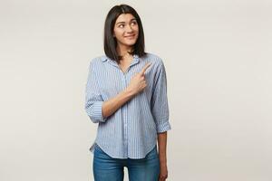 Nice young brunette looks with interest and curiosity to the right upper corner and pointing index finger there on copy space, dressed in striped shirt, isolated on a white background photo