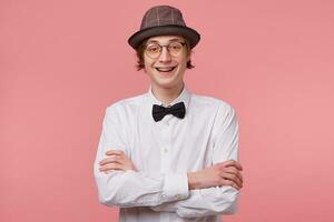Horizontal portrait of joyful nice young guy in white shirt, hat and black bowtie wears glasses happily smiling showing braces, standing with hands crossed, isolated on pink background photo