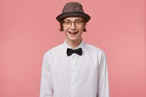 Nice young guy in white shirt, hat and black bowtie wears glasses happily widely smiling showing orthodontic brackets, isolated on pink background photo