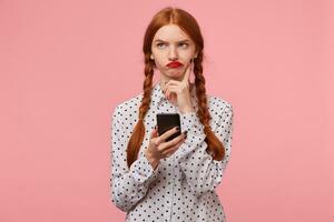 Gloomy brooding red-haired girl without enthusiasm looking in the upper left corner trying to think of what to write in a message to her friend holding the phone in her hands, on pink wall photo