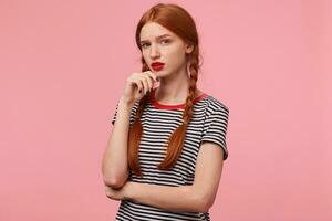 Portrait of beautiful red-haired girl with two braids keeps fist near the chin and looks sceptical, with suspect and distrust, questionably, isolated on pink wall photo