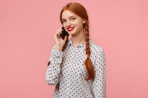 joven atractivo Pelirrojo niña con trenzas vestido en un blanco lunares camisa participación un teléfono cerca el oído con mano hablando a alguien mirando a el cámara con sonrisa,aislado en un rosado antecedentes foto