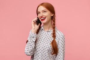 Attractive happy red-haired girl with braids dressed in a white polka-dot shirt holding a phone near the ear with hand talking to someone looking away with mouth open with laughter,isolated on a pink photo