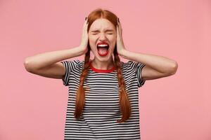 Angry young red-haired girl in stripped t-shirt, keeps closed eyes, closes her ears with palms, showing gesture of ignore, demonstrates loud scream or shout, over pink wall photo