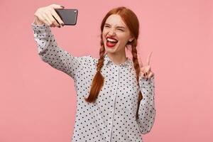 Carefree confident playful funny red-haired smiles affably, showing with her fingers a peace sign or victory, makes selfie on her phone, is wearing a white polka-dot shirt isolated on pink background photo